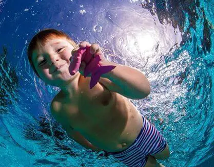 A young boy swimming in water smiling holding a pool toy confidently.