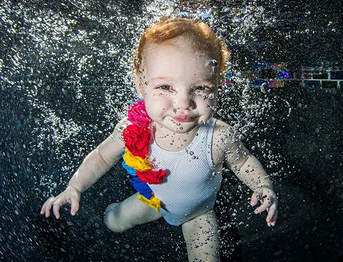 A baby in a white swimsuit with colorful ruffles swims underwater, smiling and surrounded by bubbles.
