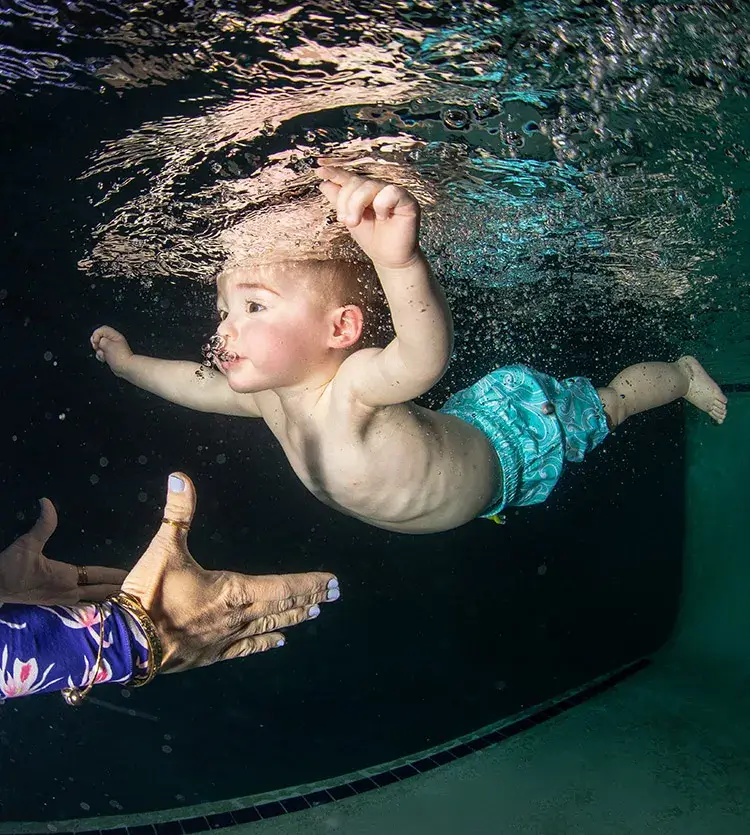 An infant in swim trunks floats underwater in a swimming pool, as an adult awaits with outstretched hands during a Gentle Submersion Workshop.