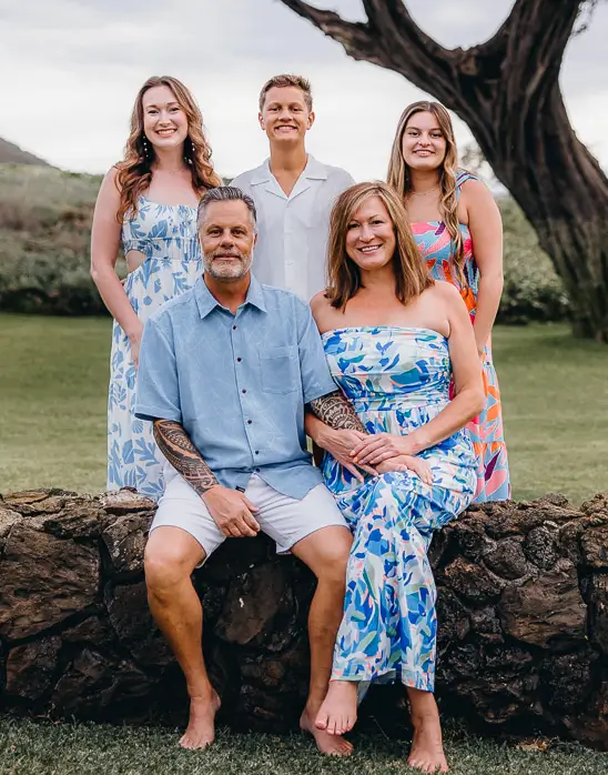 Kona's family posing outdoors in colorful summer clothing. Two adults sit on a stone wall, while three young adults stand behind them.