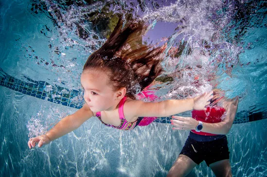 A young girl in swimsuit swims underwater in a pool with an adult supporting her.