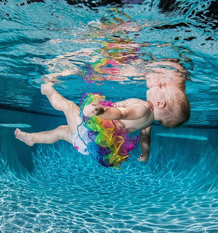 A baby wearing a colorful ruffled swimsuit floats underwater in a swimming pool, using the swim float swim method.