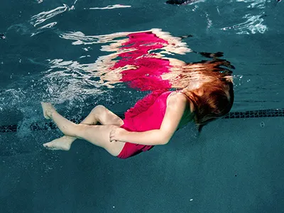 A young child in a red swimsuit floats on her back in a swimming pool, with her face just above the water, practicing the swim float swim survival swimming technique.