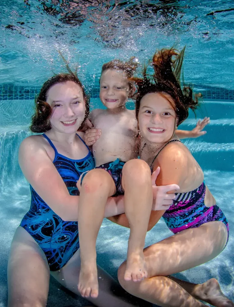 Three smiling children in swimsuits pose together underwater in a swimming pool.