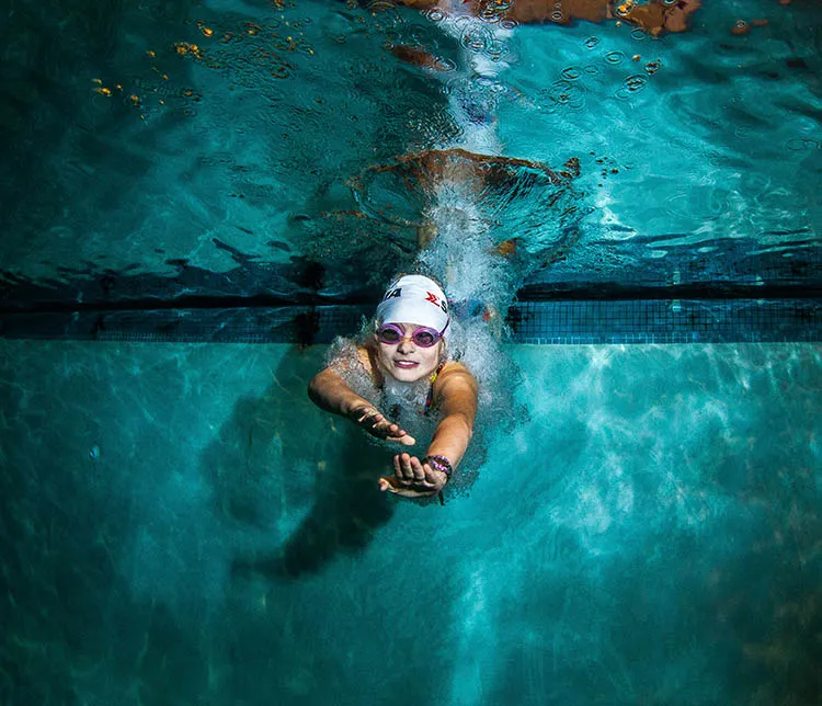 Underwater view of an adult woman swimmer wearing goggles and a swim cap, reaching forward with both hands near the edge of a swimming pool.