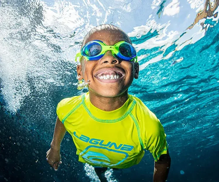 A smiling boy wearing swim goggles swims underwater, looking up toward the surface.