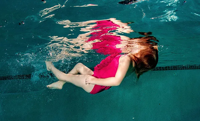 A young child in a pink swimsuit floats underwater in a swimming pool, with her face above the water's surface.